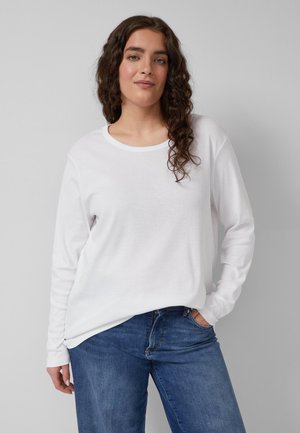 Frau mit lockigem Haar, die ein weißes Langarmshirt und blaue Jeans trägt, steht mit einer Hand in der Tasche vor einem schlichten grauen Hintergrund.