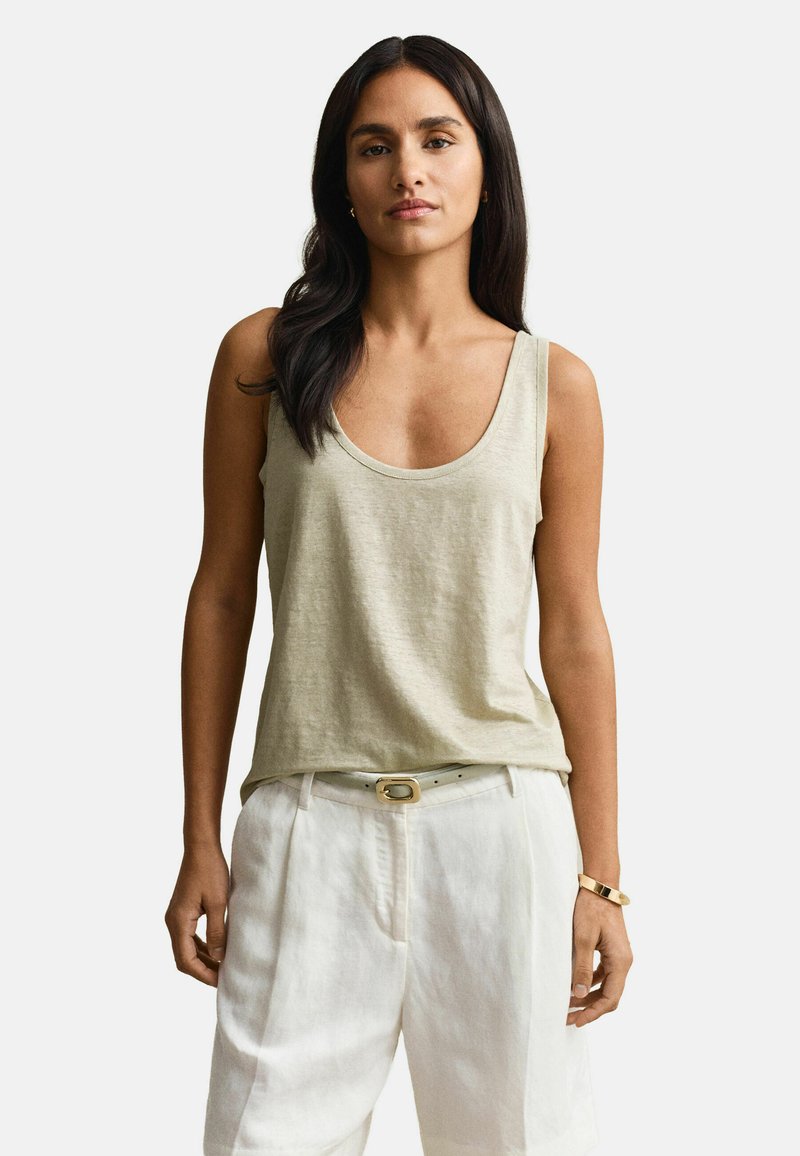 Femme aux longs cheveux foncés portant un haut beige sans manches, un short blanc avec ceinture, un bracelet en or et de petites boucles d'oreilles sur un fond uni.