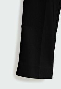 Jambe de pantalon noire avec une coupe droite et une petite fente latérale au bas de l’ourlet sur fond blanc.