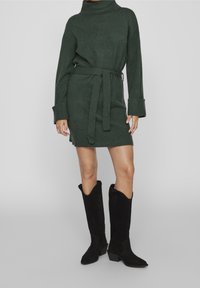 Abito lavorato a maglia verde con colletto alto e cintura da annodare. Maniche lunghe con ampi polsini. Abbinato a stivali neri in suede alti fino al ginocchio.