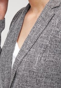 Blazer gris texturé avec un col cranté, des détails de coutures visibles et une coupe décontractée. Porté sur un haut blanc.