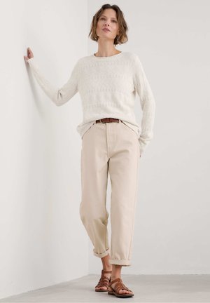 Cremefarbener Strickpullover mit einem strukturierten Rautenmuster, kombiniert mit hellbeigen hochgekrempelten Hosen und braunen Sandalen mit Riemen. Wand im Hintergrund.