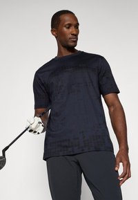 Námornícka polo košeľa s krátkym rukávom a jemným geometrickým vzorom, vyrobená z ľahkého materiálu. Model drží golfovú palicu a má na sebe rukavicu.