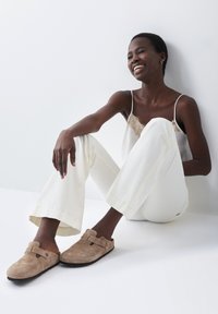 Zuecos de ante marrón con una correa de hebilla, combinados con pantalones de pierna ancha color crema y una blusa ligera con detalles de encaje, sobre un fondo liso.