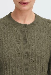 Vihreä neulecardigan, jossa on teksturoitu kuvio, pyöreä pääntie ja kuusi tummaa nappia edessä. Kangas vaikuttaa pehmeältä.