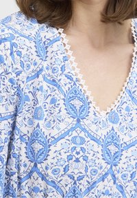 Modrý a bílý květinový vzorovaný textil s výstřihem ve tvaru V a bílou krajkou okolo výstřihu, zobrazující složité botanické vzory.