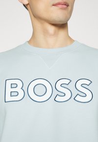Ljusblå sweatshirt med rund halslinning och broderad "BOSS"-logotyp i vitt med mörkblå detaljer. Mjuk, texturerad tyg.
