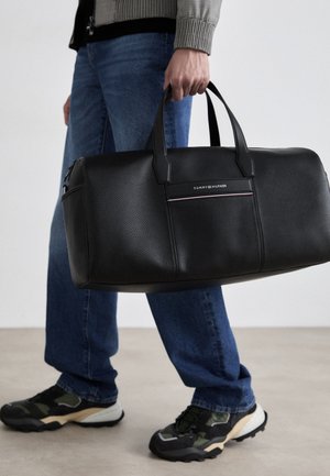 Zwarte leren reistas met twee bovenhandvatten, met een strak ontwerp en een logo-accent. Robuuste textuur en zichtbare ritsluiting.