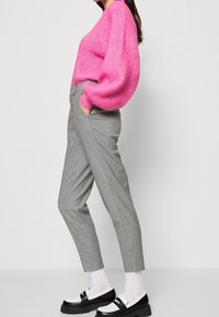 Femme portant un pull rose vif, un pantalon à carreaux slim-fit, des chaussettes blanches et des mocassins noir et blanc, se tenant devant un fond uni.