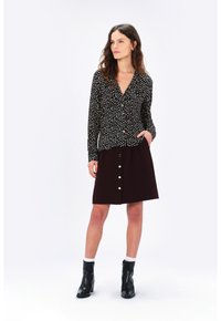 Chemise noire à manches longues avec des pois blancs et rouges, associée à une jupe évasée bordeaux comportant des boutons blancs sur le devant. Bottines noires.