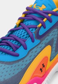 Κοντινό πλάνο μπλε αθλητικού παπουτσιού με μοβ κορδόνια, κίτρινα και πορτοκαλί στοιχεία, ύφασμα από δίχτυ και το λογότυπο Under Armour στη γλώσσα και στην πλευρά.