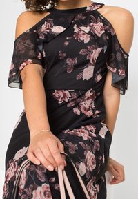 Svart blommig klänning med cut-out axlar, genomskinliga volangärmar och ros-mönster i nyanser av rosa och kräm på ett slätt tyg.