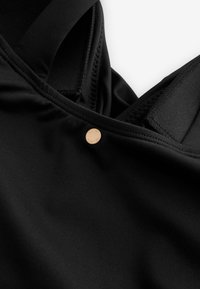 Prim-plan al unui articol vestimentar din țesătură neagră, cu bretele late și un nasture mic, rotund, aurit, pe cusătură lângă decolteu.