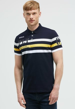 Mann trägt ein marineblaues Poloshirt mit weißen und gelben horizontalen Streifen und Text, steht vor einem schlichten hellen Hintergrund.