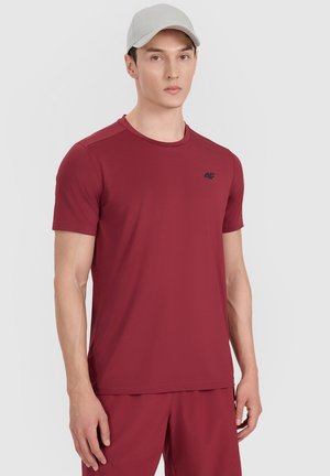 Joven con camiseta deportiva de manga corta burdeos y pantalones cortos a juego con una gorra de béisbol gris claro, de pie frente a un fondo liso.