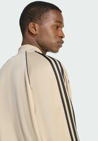 Béžová športová bunda s vysokým golierom, s čiernymi vertikálnymi pruhmi pozdĺž ramien. Hladká textília s jemnou štruktúrou.