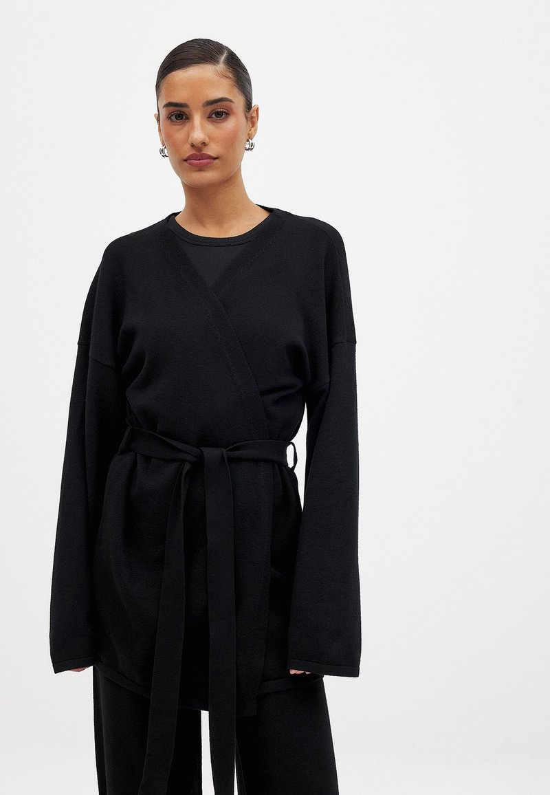 Juodas apvyniotas kardiganas, pagamintas iš minkštos medžiagos, su ilgomis rankovėmis, dirželiu ir laisvu siluetu. Lygus paviršius, minimalus dizainas.