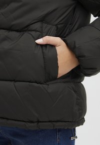 Juodas pūkinis švarkas su siūtomis sekcijomis, užsegama kišene ir reguliuojamu apvadu. Medžiaga atrodo lygi ir izoliuota.