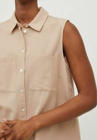 Chemise sans manches en lin beige avec un col pointu, deux poches poitrine et fermeture à boutons sur le devant. Texture lisse avec une coupe décontractée.