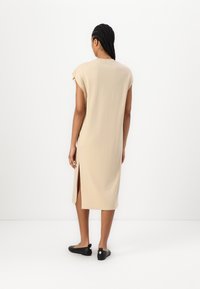 Robe midi beige avec manches courtes, fente latérale, texture de tissu douce et coupe décontractée, associée à des chaussures plates noires, vue de dos.