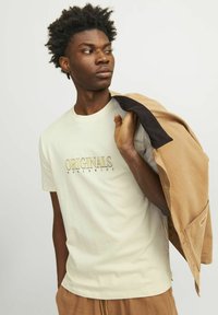 Camiseta de color crema claro con "ORIGINALS WORLDWIDE" impreso en dorado. El modelo sostiene una chaqueta marrón sobre su hombro, con pantalones marrones visibles.