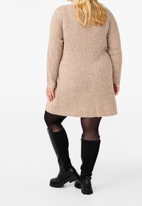 Beiges, geripptes Strickkleid mit langen Ärmeln, schwingendem Saum, kombiniert mit knielangen schwarzen Stiefeln und schwarzen Strumpfhosen. Texturierte Stoffdetails sind sichtbar.