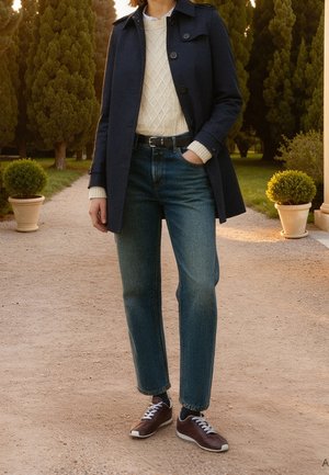 Person trägt einen marineblauen Mantel, weißen Strickpullover, blaue Jeans und braune Sneakers und steht auf einem Kiesweg mit Topfpflanzen und hohen Bäumen in der Nähe.