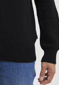 Nahaufnahme einer Person, die einen schwarzen strukturierten Strickpullover mit gerippten Bündchen und Saum trägt, kombiniert mit blauen Jeans.