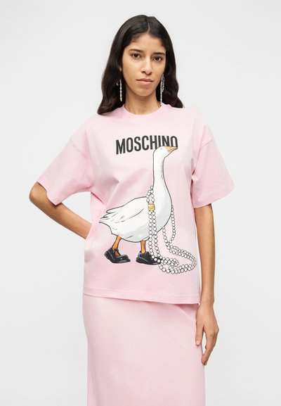 Ροζ βαμβακερό μπλουζάκι με μια εικόνα χήνας που φοράει μαύρα παπούτσια και μαργαριτάρια, με τη λέξη «MOSCHINO» τυπωμένη επάνω σε έντονα μαύρα γράμματα.