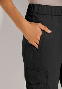 Hand met een lichte huidtint rustend in de voorkeurszak van zwarte cargobroek met elastische tailleband en extra zijzakflap.