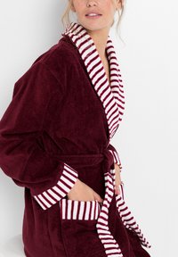 Robe in velluto bordeaux con una texture morbida, caratterizzato da bordi a strisce bianchi e bordeaux sul colletto sciallato e sulle maniche, e tasche frontali.