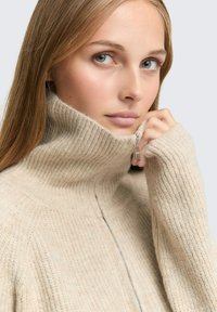 Beżowy sweter z prążkowanego dzianiny z wysokim kołnierzem i zamkiem błyskawicznym na pół, wykonany z miękkiej, teksturowanej tkaniny o luźnym kroju.