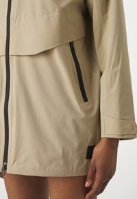 Veste beige à manches longues avec détails de fermeture éclair noirs et poche à rabat, portée par une personne avec main et haut de la cuisse visibles.