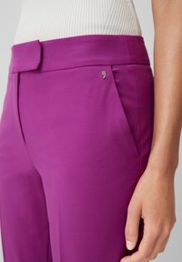 Pantalon ajusté en tissu violet vif, avec une ceinture plate, des poches latérales et un petit accent argenté à l'avant. Surface texturée.