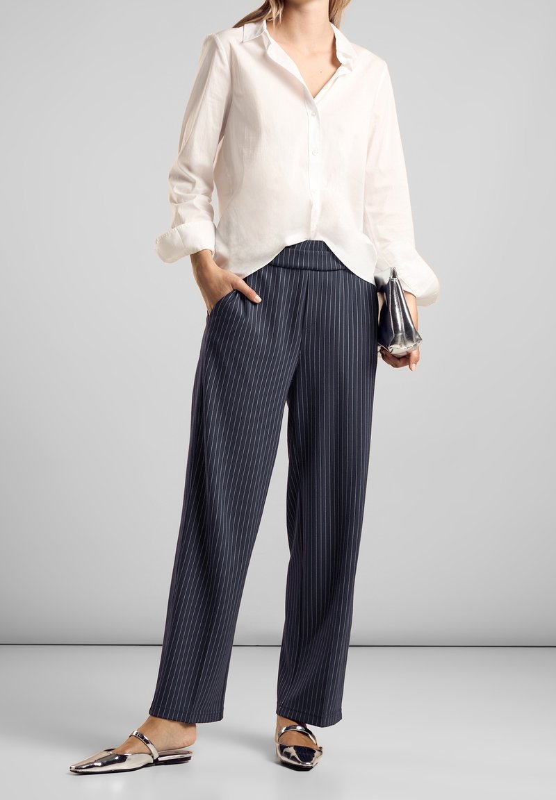 Chemise en lin blanc à boutons avec les manches retroussées ; pantalon taille haute à rayures verticales bleu marine ; ballerines métalliques à bout pointu ; petite pochette en argent.