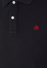 Polo noir en tissu texturé, avec trois boutons blancs sur le col et un petit logo rouge brodé sur la poitrine.