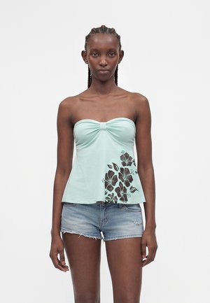 Jeune femme aux cheveux tressés portant un top bustier vert menthe avec un motif floral noir et un short en jean bleu clair.
