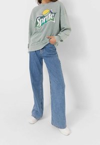 Jasnozielona bluza z dużym logo Sprite, połączona z szerokimi niebieskimi dżinsami i białymi sneakersami. Casualowy, luźny strój.