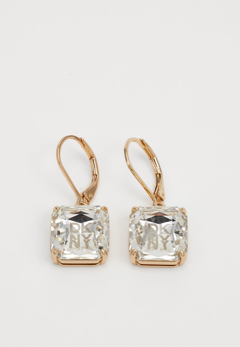 Brincos de alavanca em ouro com grandes cristais quadrados claros gravados com o logótipo "DKNY" em fundo branco.
