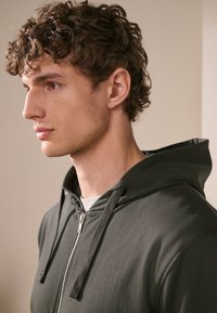 Donkergroene zip-up hoodie met een gladde textuur, voorzien van een trekkoordkap en een zilveren rits. Eenvoudig, minimalistisch ontwerp zonder patronen.
