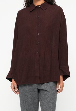 Femme portant une chemise ample marron foncé à boutons avec manches longues et un pantalon gris sur un fond uni.