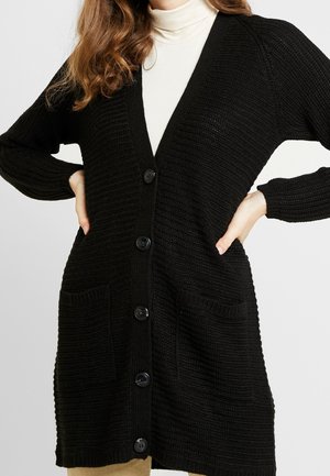 Femme portant un long cardigan en maille noir avec des poches avant et de gros boutons, par-dessus un pull à col roulé blanc.