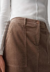 Gonna di velluto a coste marrone con texture rigata verticale, dotata di una tasca anteriore e una chiusura con un singolo bottone, abbinata a una maglietta bianca a maniche lunghe.