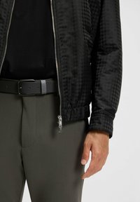 Schwarze Bomberjacke mit Hahnentrittmuster, metallischem Reißverschluss und elastischen Bündchen, getragen über einem schwarzen Hemd und grauen Hosen mit einem schwarzen Gürtel.