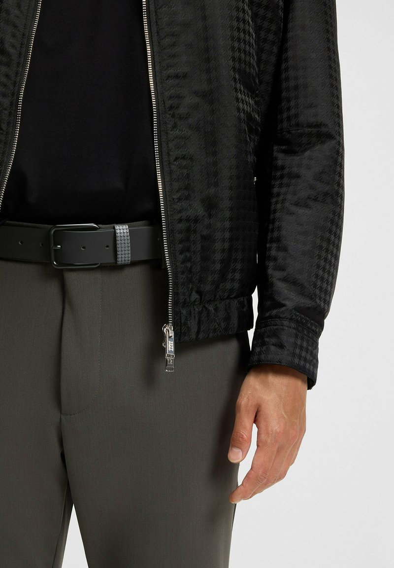 Schwarze Bomberjacke mit Hahnentrittmuster, metallischem Reißverschluss und elastischen Bündchen, getragen über einem schwarzen Hemd und grauen Hosen mit einem schwarzen Gürtel.