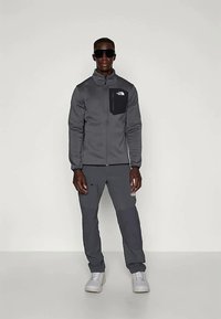 Grå zip-up jacka med en svart bröstficka, matchande grå byxor och vita sneakers. Slät tygtextur, figurnära design och minimal varumärkeslogg.