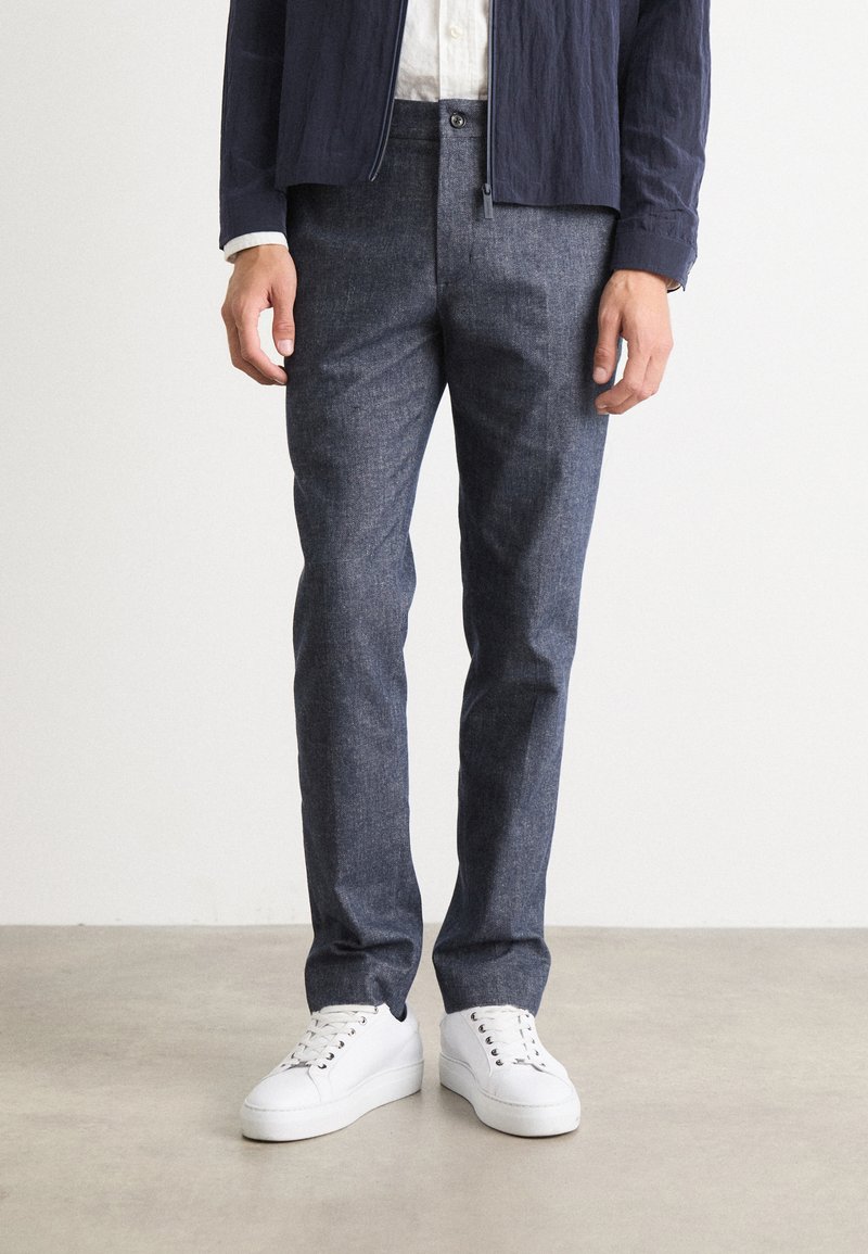 Pantalon en denim bleu à coupe slim, avec un tissu texturé et une fermeture à bouton, associé à des baskets blanches.