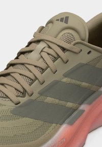 Κοντινό πλάνο ενός μπεζ και ελιάς αθλητικού παπουτσιού Adidas με ύφασμα από δίχτυ, τρεις σκούρες ρίγες, μπεζ κορδόνια και ροζ μαλακή σόλα.