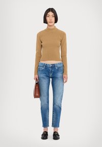 Pull col roulé côtelé marron clair, jean taille haute bleu, mocassins noirs avec des accents dorés, tenant un petit sac à main marron.