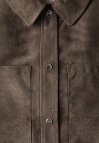 Braunes Cordhemd mit Kragen, silbernen Druckknöpfen und zwei Vordertaschen mit vertikaler Rippstruktur.